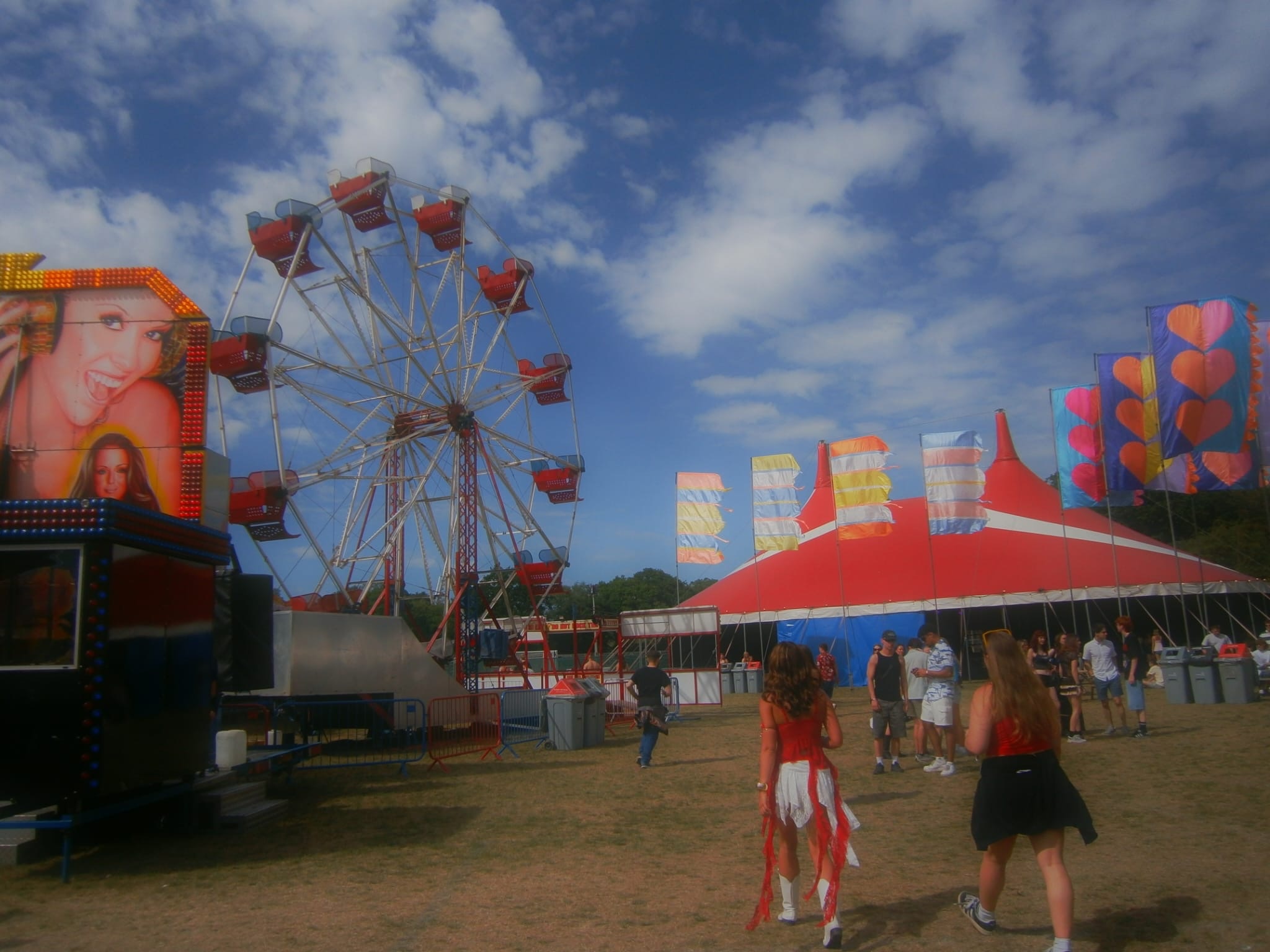 A Ferris wheel and a red-and-white circus tent at a fairground, with people walking across the grass under a partly cloudy sky.