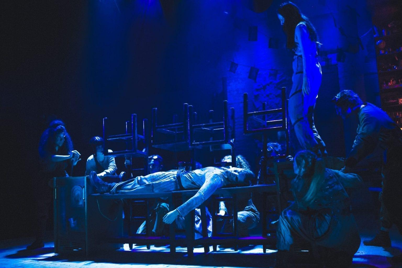 BA (Hons) Acting students in the middle of a performance of Frankenstein. The actors are gathered around a table in the middle of the stage, on which another actor is lying.