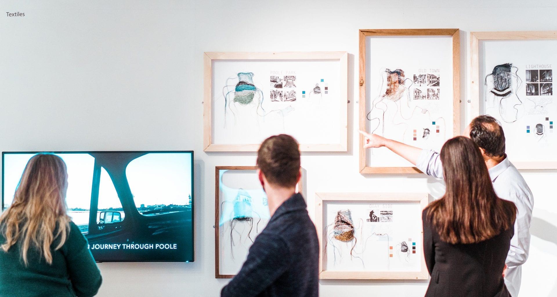 People study several framed pictures that are hanging on a gallery wall. Another person watches something on a television screen next to them.