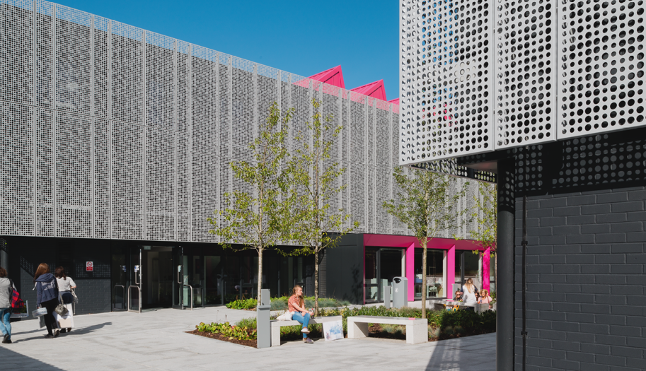 A large university building with metal mesh exteriors, pink square arch doorways and a pink spike roof. There is a courtyard with trees and benches outside.