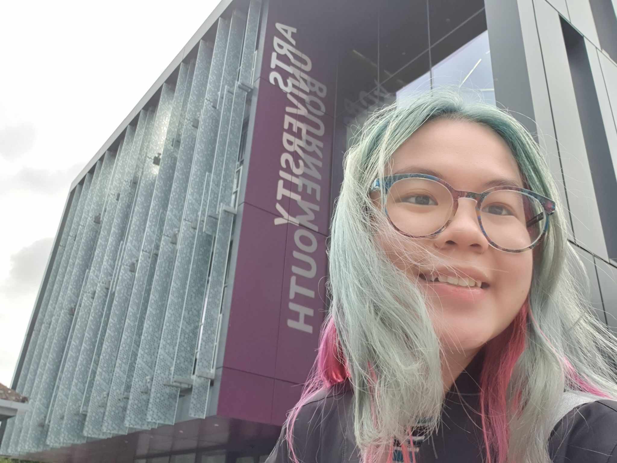 Ngan Nguyen taking a selfie outside AUB North Building's purple banner, with turquoise hair and glasses.