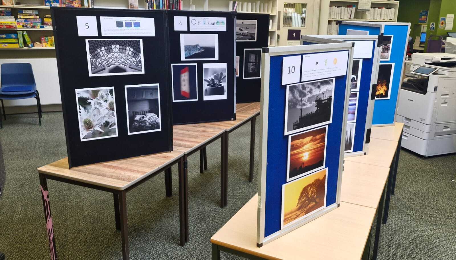 Display boards with mounted photographs on wooden tables in library setting with bookshelves and blue chairs visible in background.