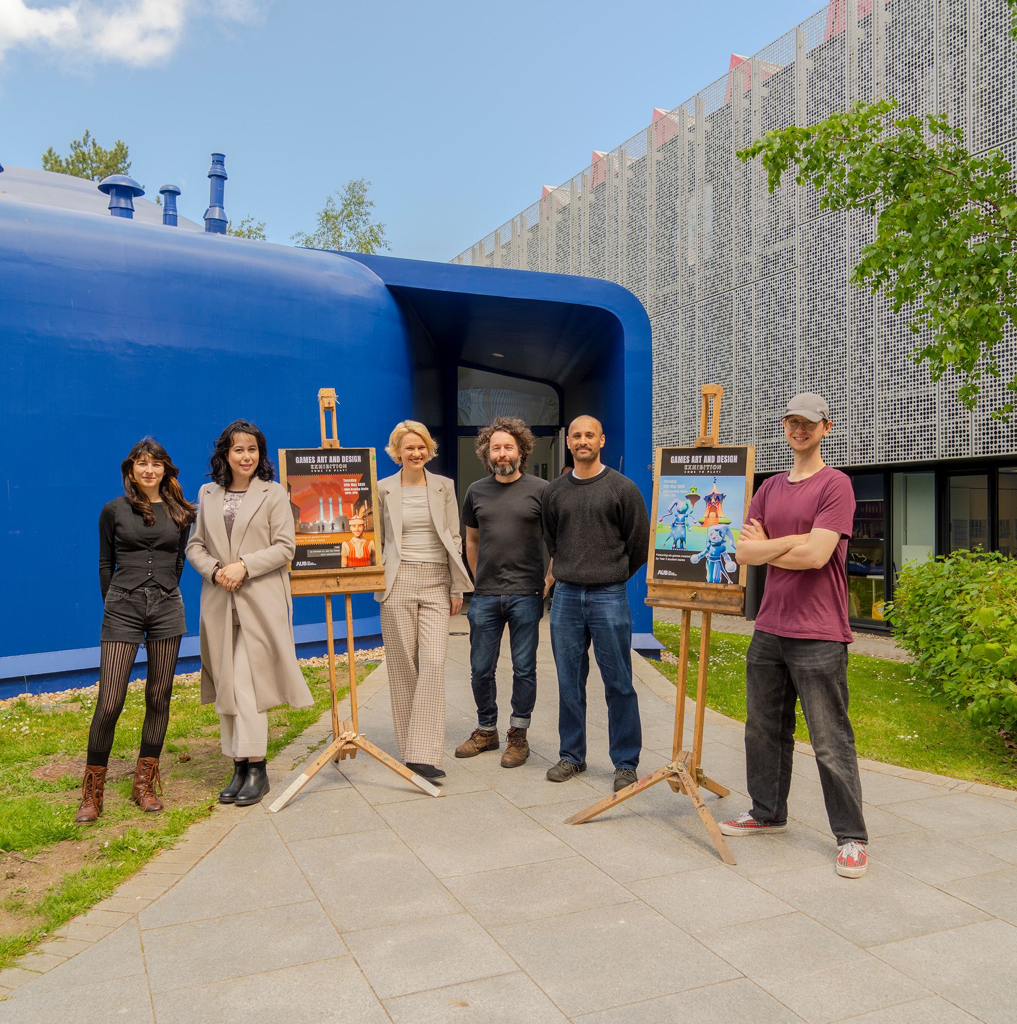 Several members of Arts University Bournemouth BA (Hons) Games Art and Design staff stood outside the blue CRAB Drawing Studio on campus with two posters on easels posing for a photo.