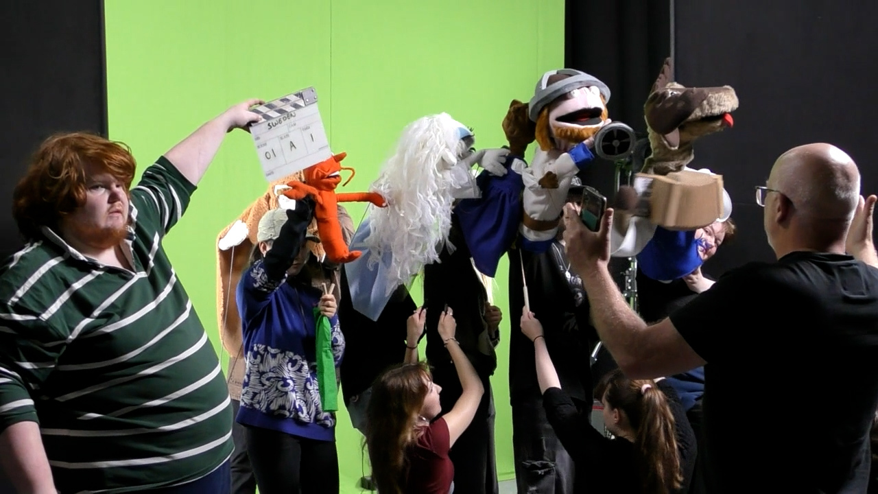 BA (Hons) Modelmaking students holding assorted puppets while a member of technical staff holds a clapper board next to them, all standing in front of a greenscreen in a studio at Arts University Bournemouth.
