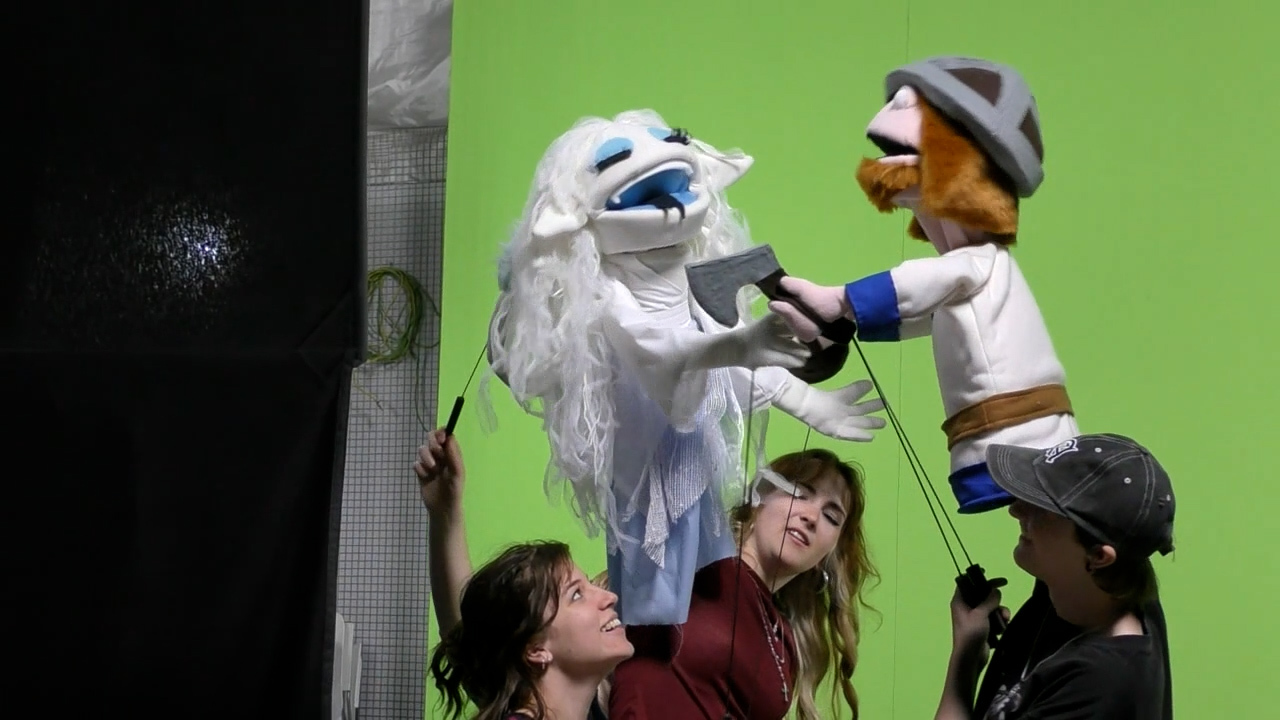 BA (Hons) Modelmaking students holding two puppets, one in the style of an ice queen, the other in the style of a Viking, performing in front of a greenscreen at Arts University Bournemouth.