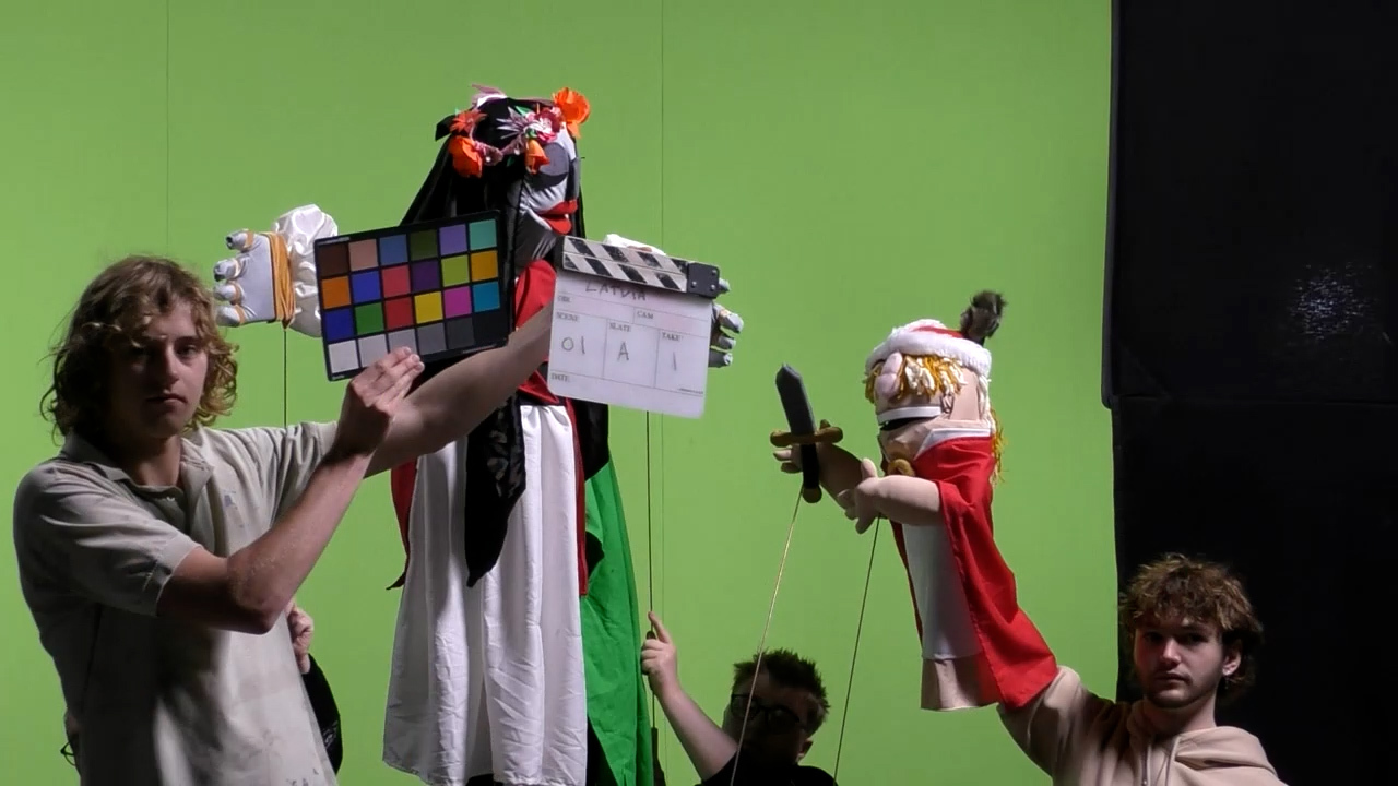 A student holds a clapper board and a colour grading chart in front of a tall white female-style puppet. Another student holds a smaller puppet wearing a red robe and holding a sword.