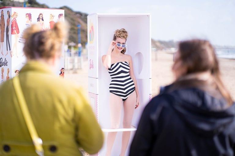 A model in a black and white swimsuit dressed as a Barbie doll standing in a rectangular box on a beach peers over her sunglasses.