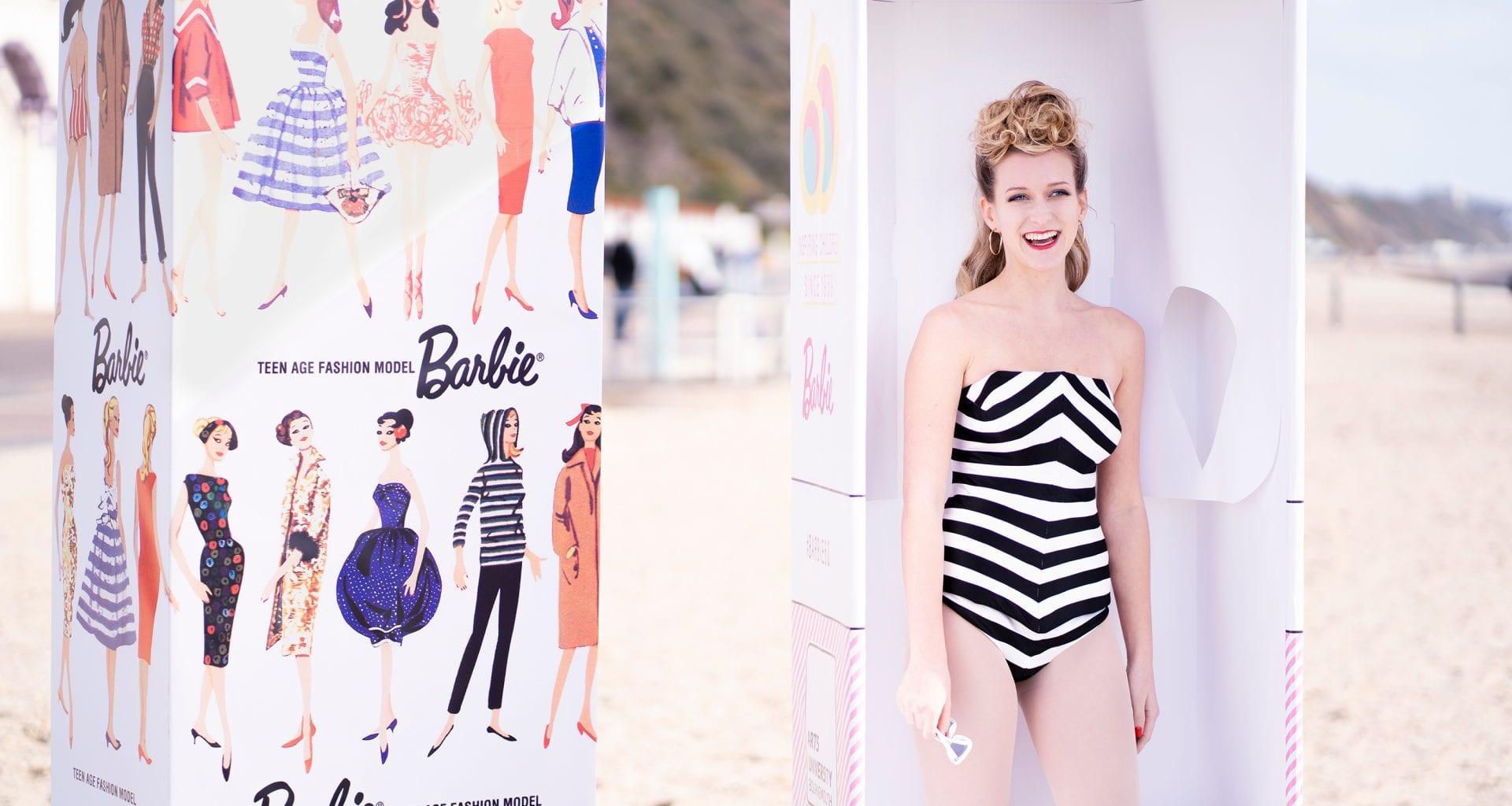 Closer photo of a model in a black and white swimsuit poses as a Barbie doll in a rectangular box on a beach. Another box with a selection of Barbie artworks stands next to them.