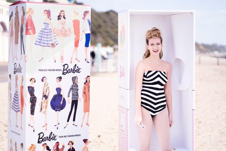 A model in a black and white swimsuit poses as a Barbie doll in a rectangular box on a beach. Another box with a selection of Barbie artworks stands next to them.