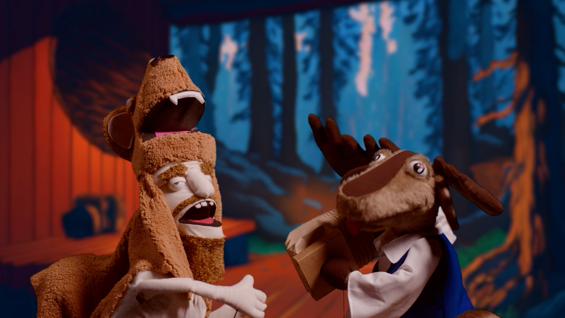 A puppet of a bearded man wearing a wolf's head headdress and a puppet of a moose, with a blue, winter forest scene in the background.