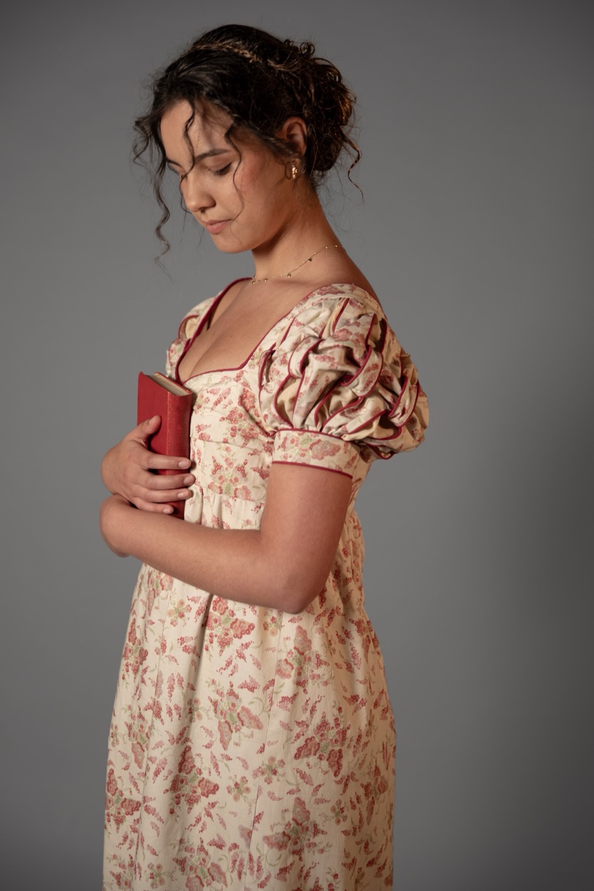 A young woman in a floral dress holds a red book against a grey studio backdrop.