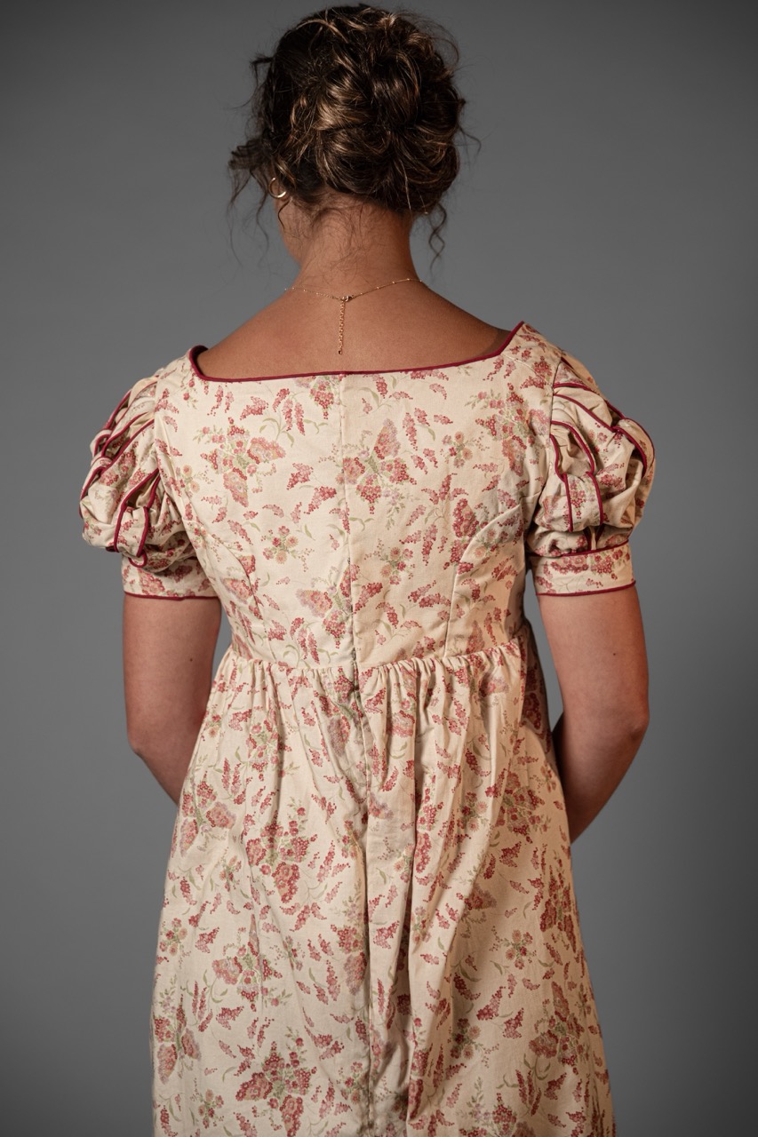 A woman seen from behind wearing a cream floral dress with puff sleeves, standing against a plain grey background.