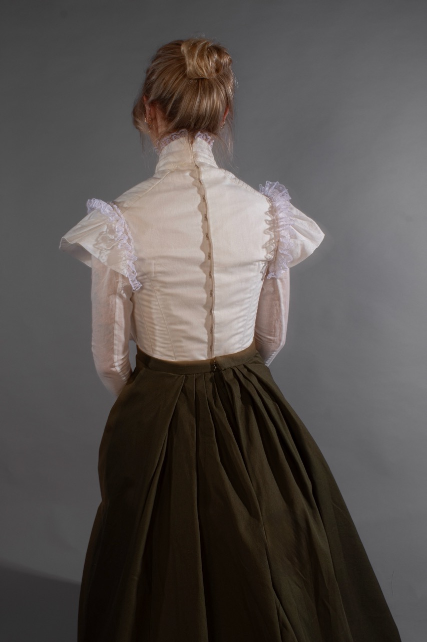 A woman seen from behind wearing a sheer cream blouse with ruffled shoulders and a dark full skirt, posed against a plain grey background.