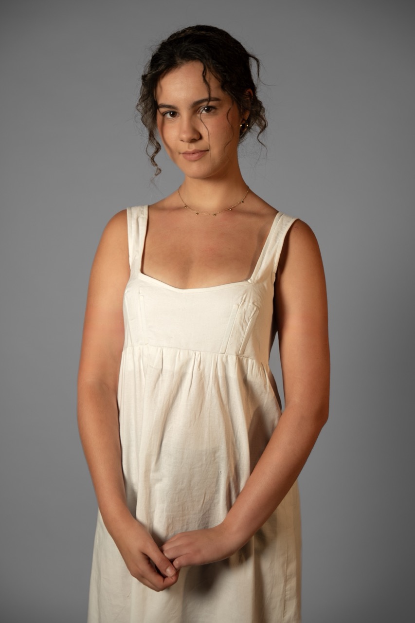 A young woman in a cream sleeveless dress stands against a plain grey background.