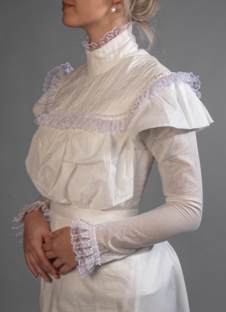 A woman in a white high-neck dress with lace trim stands against a plain grey background.