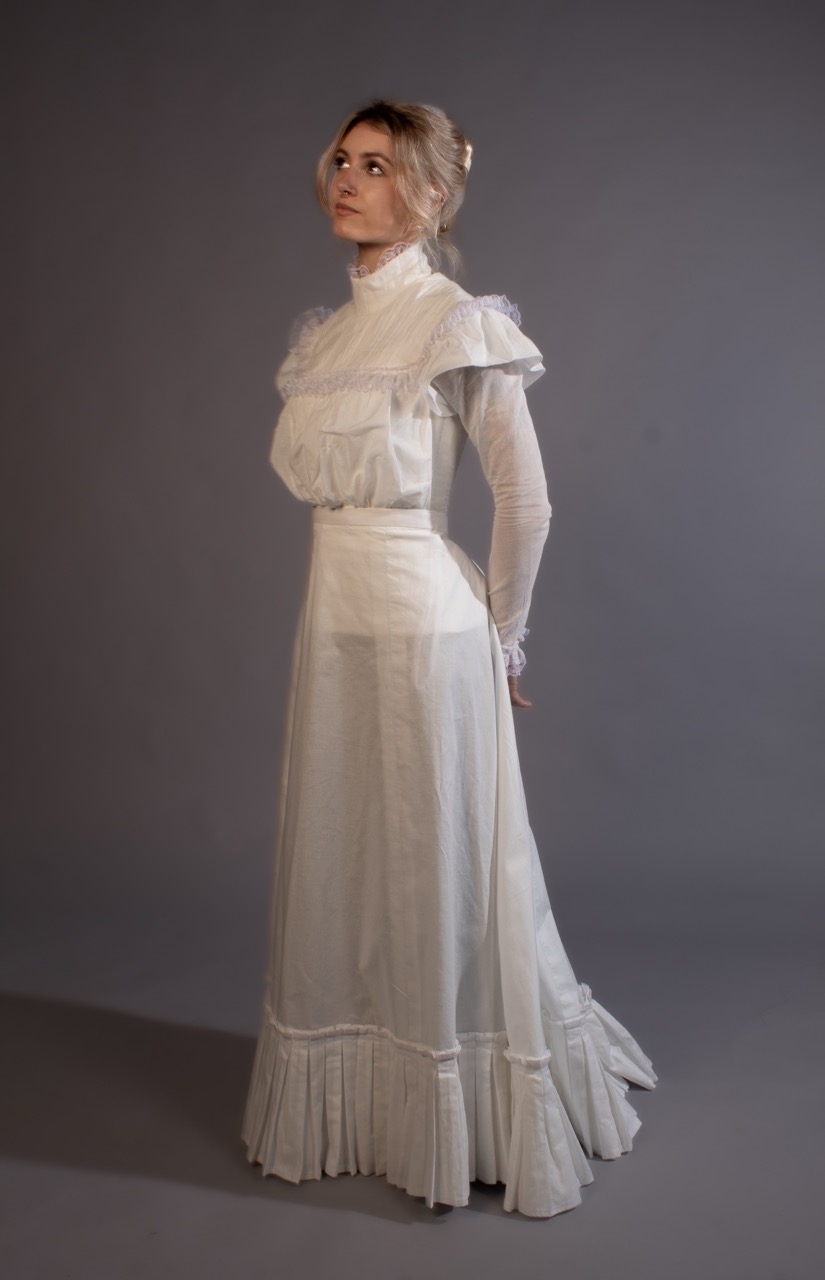 A woman in a white vintage-style dress stands against a plain grey studio backdrop.