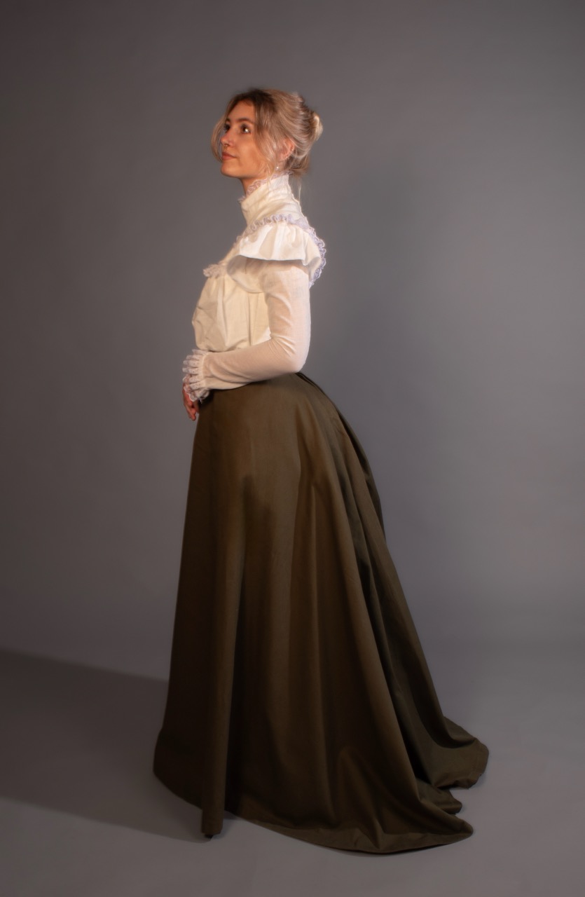 A woman in a white ruffled blouse and long dark skirt stands in profile against a plain grey studio backdrop.