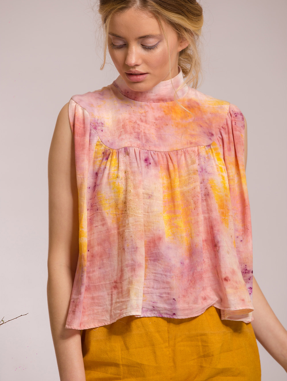 Woman wearing sleeveless tie-dye top in pink, orange and yellow tones with high neckline, paired with mustard yellow trousers.