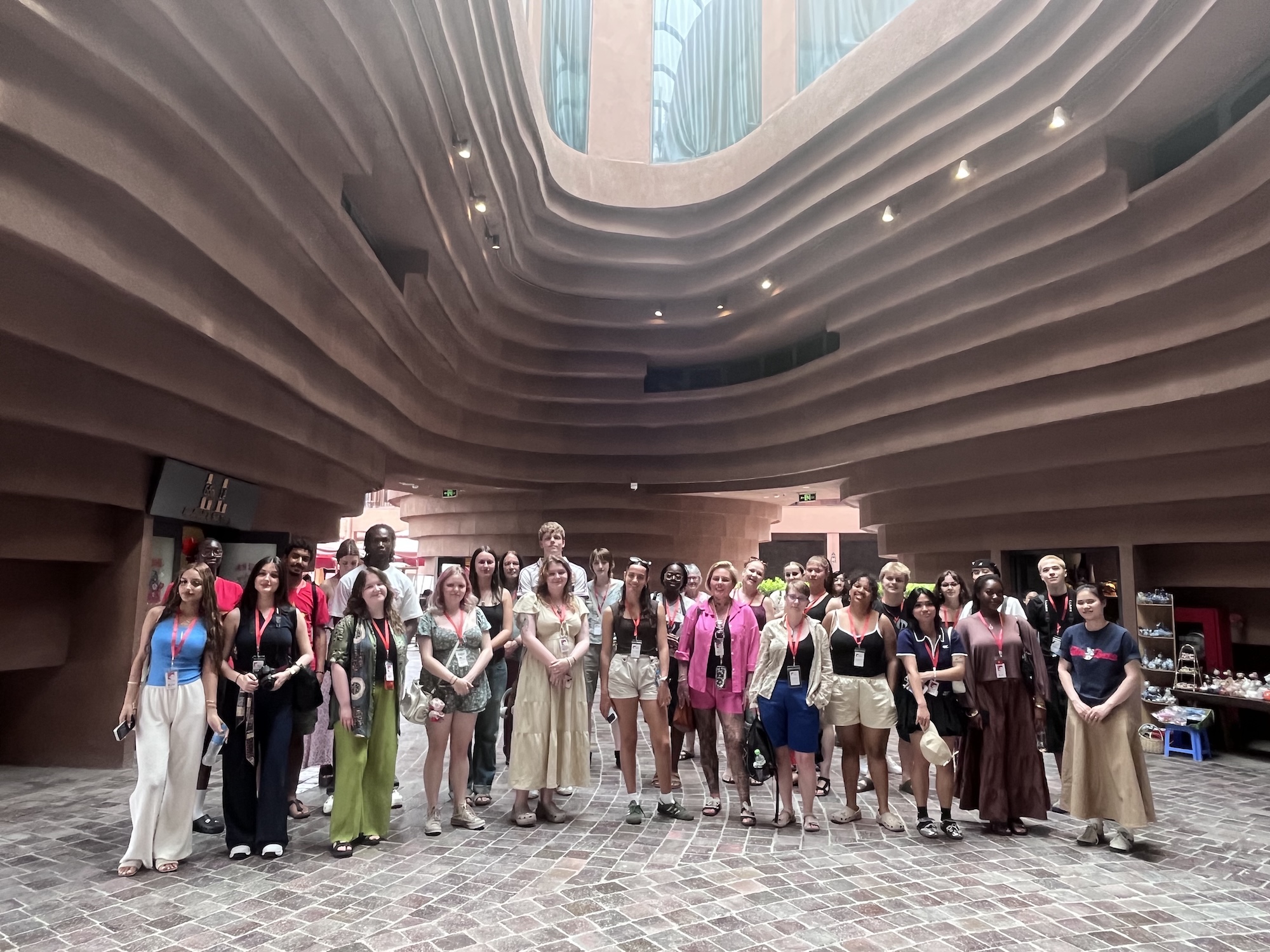 A group of approximately 30 people stands together in a spacious, modern interior with curved brown walls and large windows. The group features a diverse mix of individuals, dressed in various colourful outfits, arranged in two rows, facing the camera.