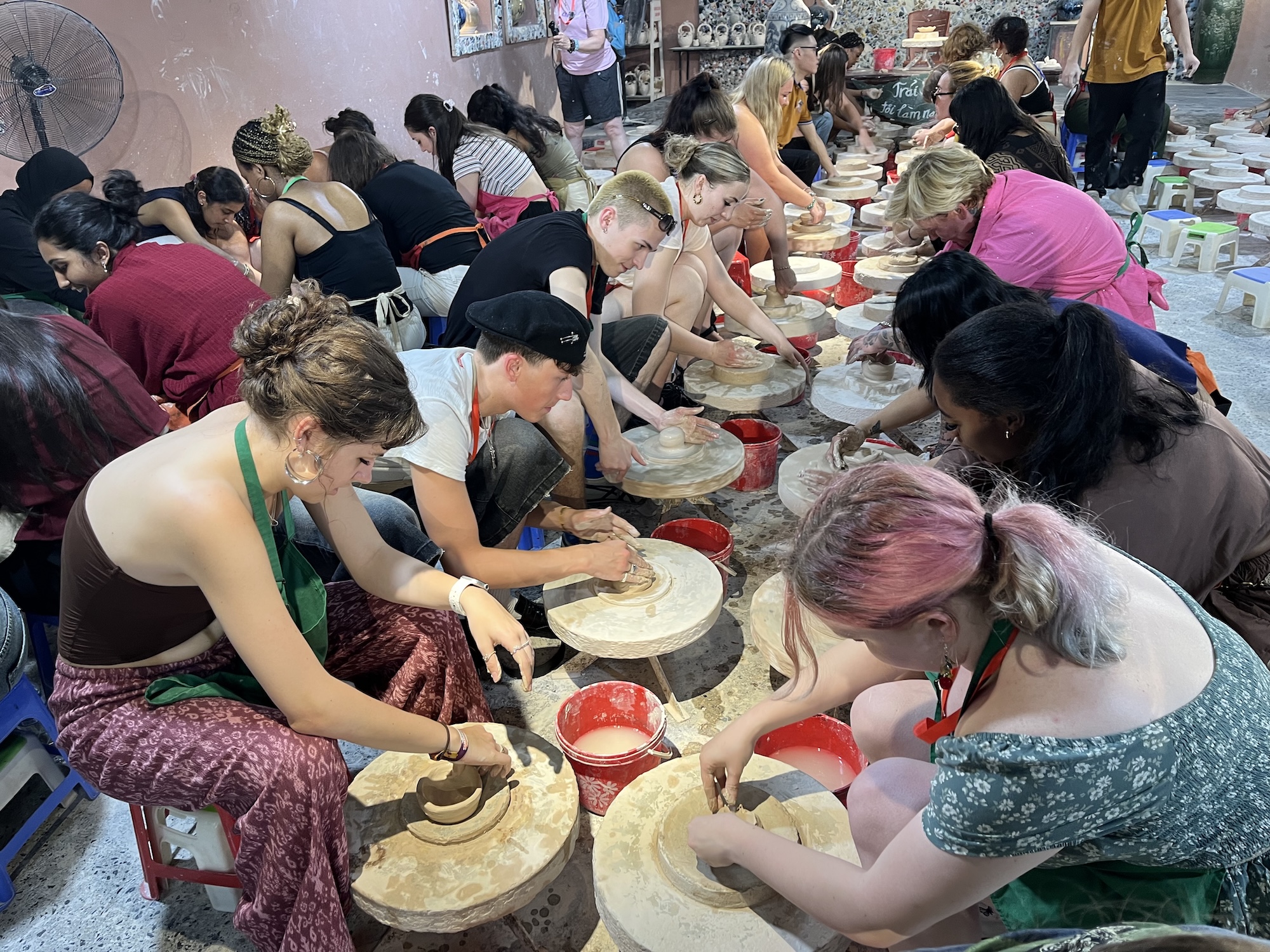 A group of individuals engaged in pottery-making, seated at pottery wheels in a workshop. Each person is concentrating on shaping clay, with tools and bowls for collecting excess clay visible on the ground.