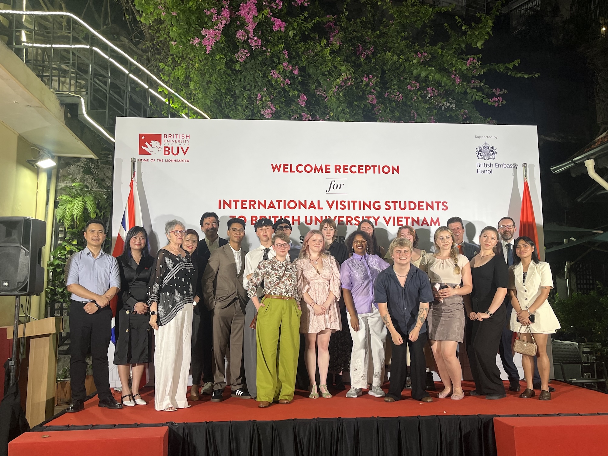 A group of diverse students pose for a photograph in front of a large banner that reads "Welcome Reception for International Visiting Students at British University Vietnam." The banner is flanked by flags. The setting is outdoor with greenery.