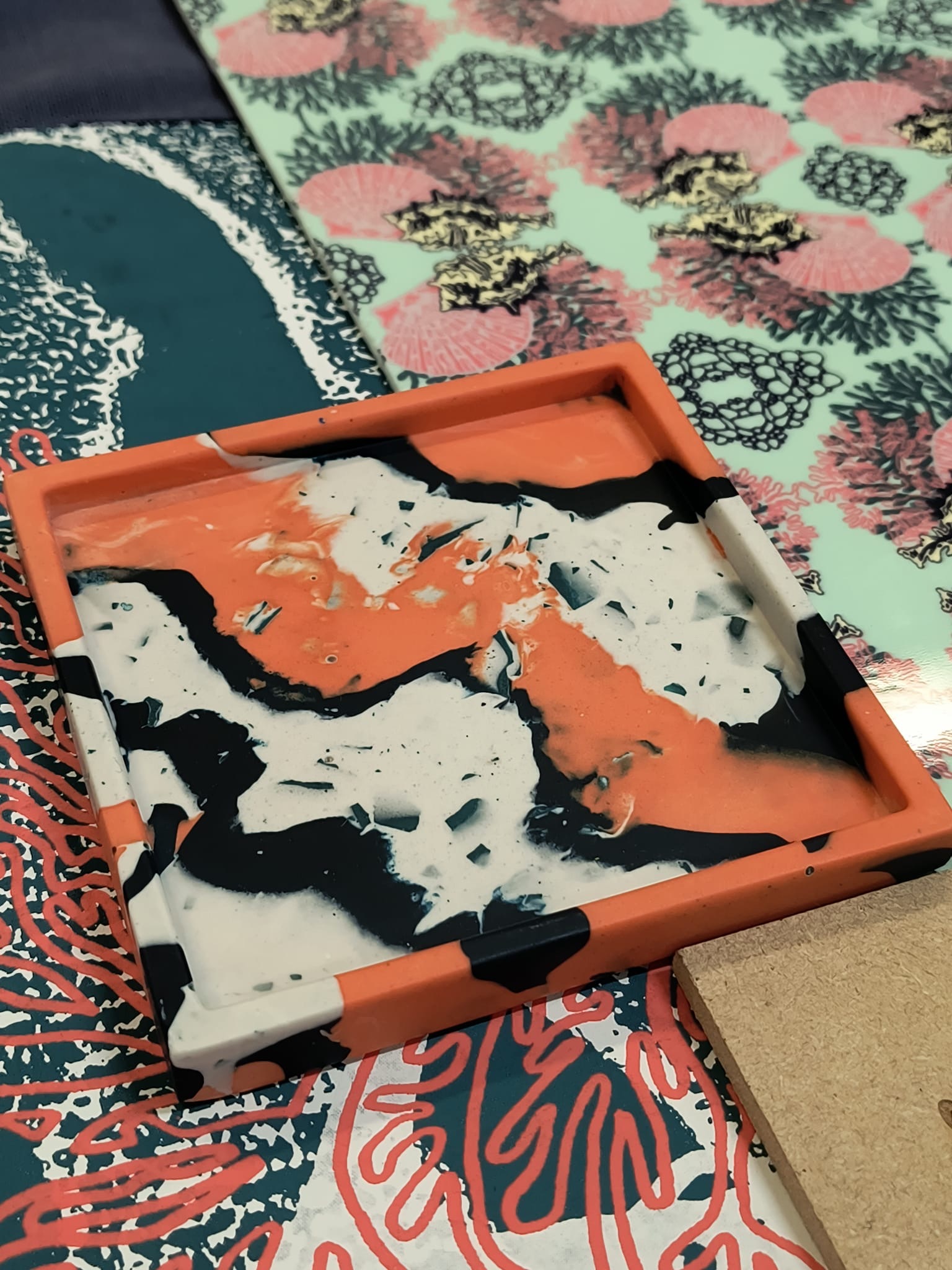 Orange-framed screen printing frame with black and white ink residue on mesh, positioned over colourful floral patterned fabric.