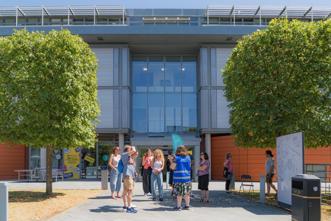 Modern glass-fronted building with blue facade, flanked by two large green trees. Group of people gathered on paved courtyard area.