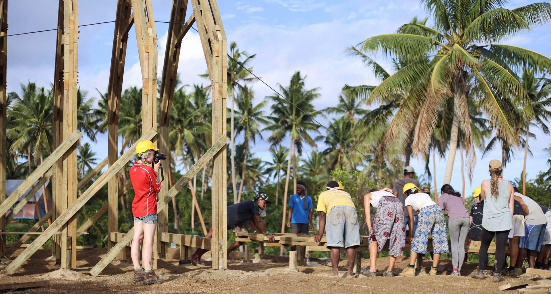 A group of people work on a wooden base for a building. A photographer stands to the left and takes pictures. There are palm trees in the background.