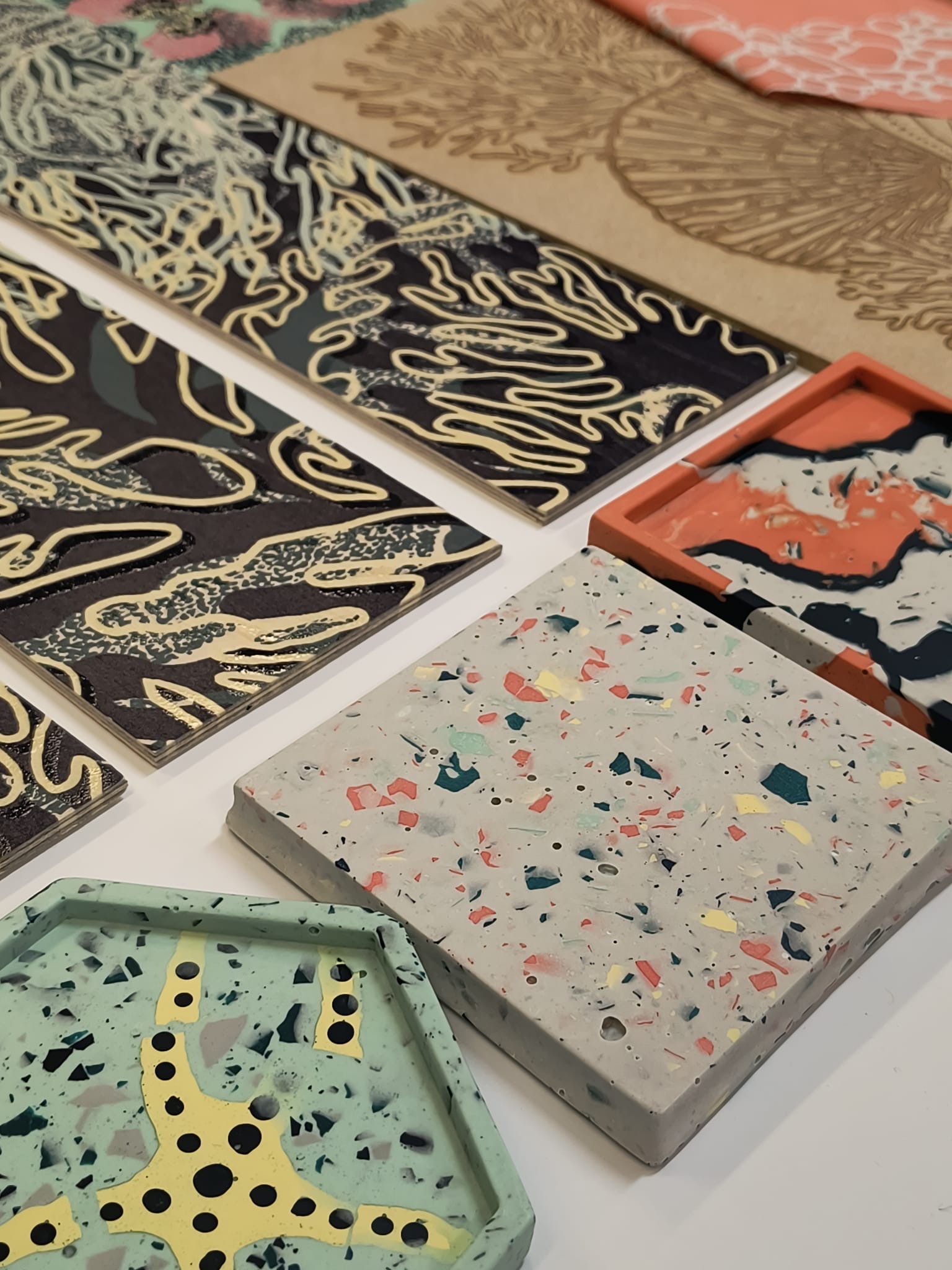 Various decorative tiles and samples arranged on white surface, featuring terrazzo patterns, marbled designs in pink and coral, and black tiles with gold flourishes.