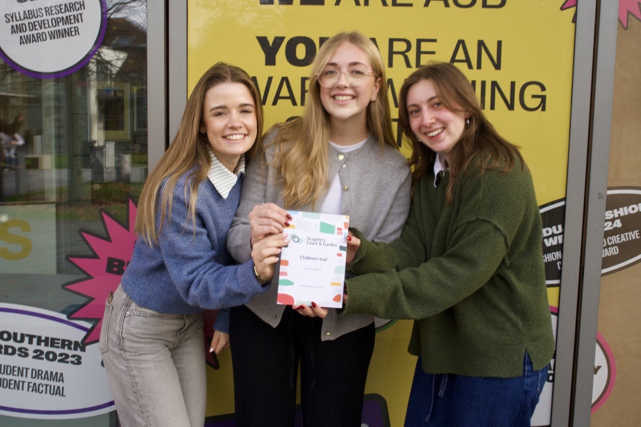 Three young women holding a certificate, smiling at camera. Yellow banner with black text visible behind them. School uniform and casual clothing.