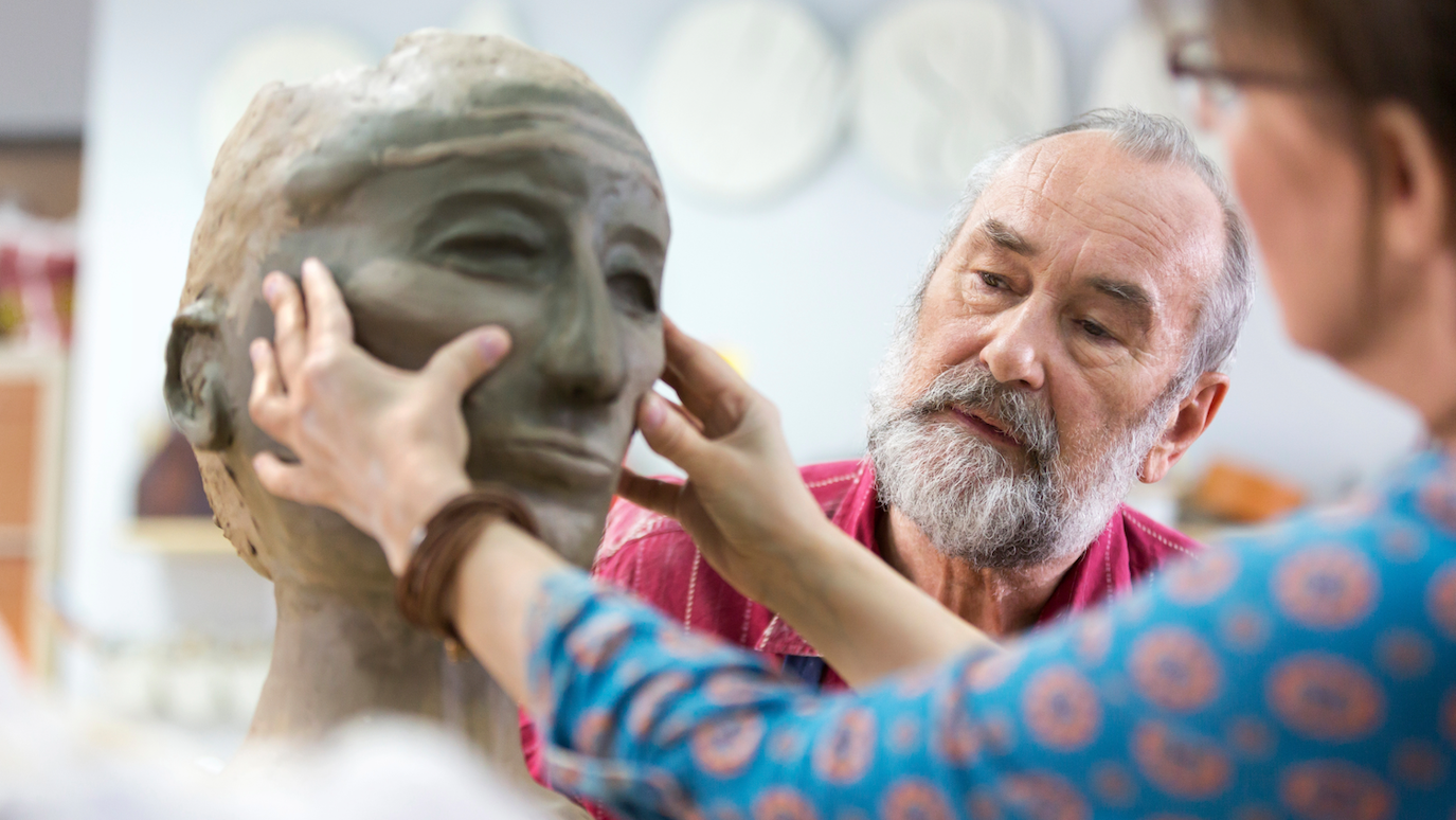Bearded man in red shirt sculpting clay head whilst woman in patterned blue top assists, hands positioned on clay sculpture