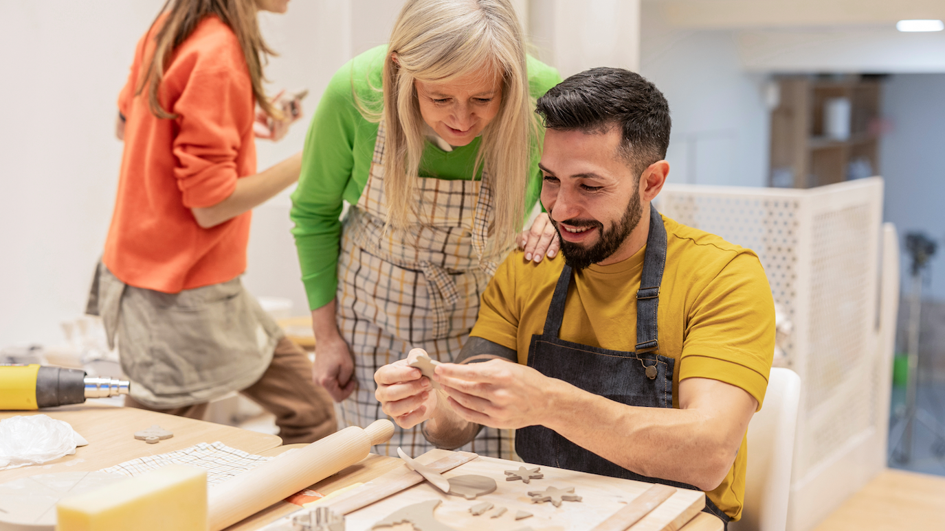 Man in yellow shirt and apron working with clay on pottery wheel whilst woman in green top and apron assists, with another person in orange top nearby