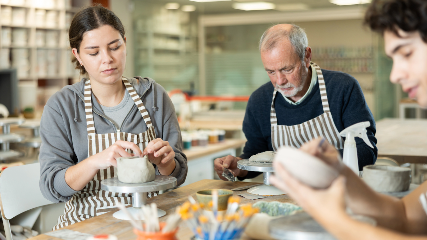 Three people in striped aprons working with clay pottery in a bright workshop with shelves of ceramic pieces in background.