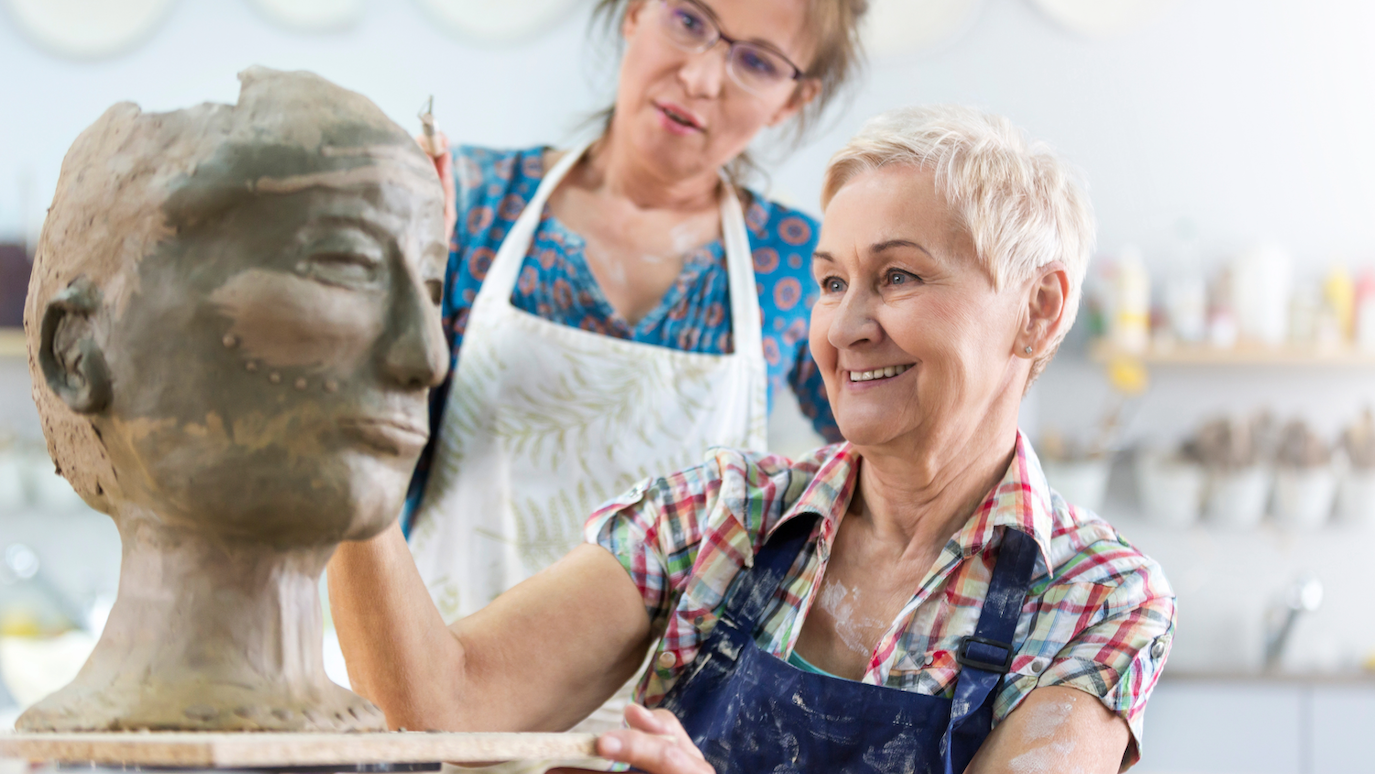 Two women in aprons working on clay sculpture of human head in pottery studio
