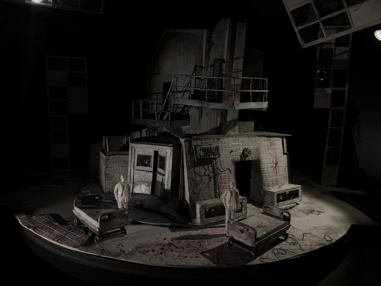 A damaged stage set resembling a multi-storey building with railings and windows, surrounded by overturned beds and mannequins under dim lighting.