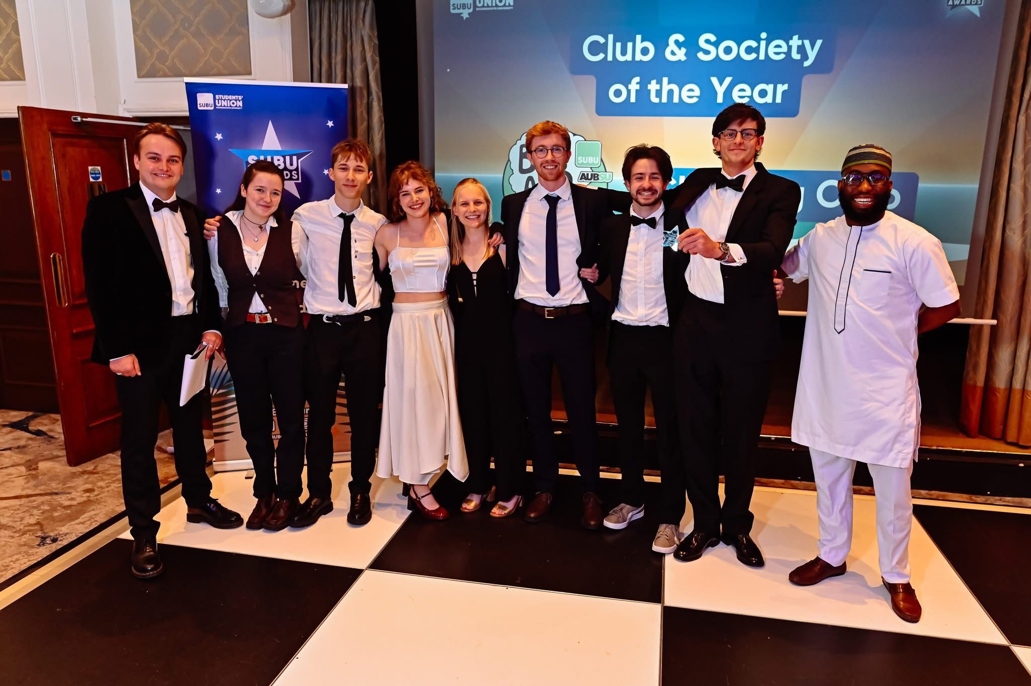 Group of ten people in formal attire posing at awards ceremony, blue screen displays "Club & Society of the Year" behind them.