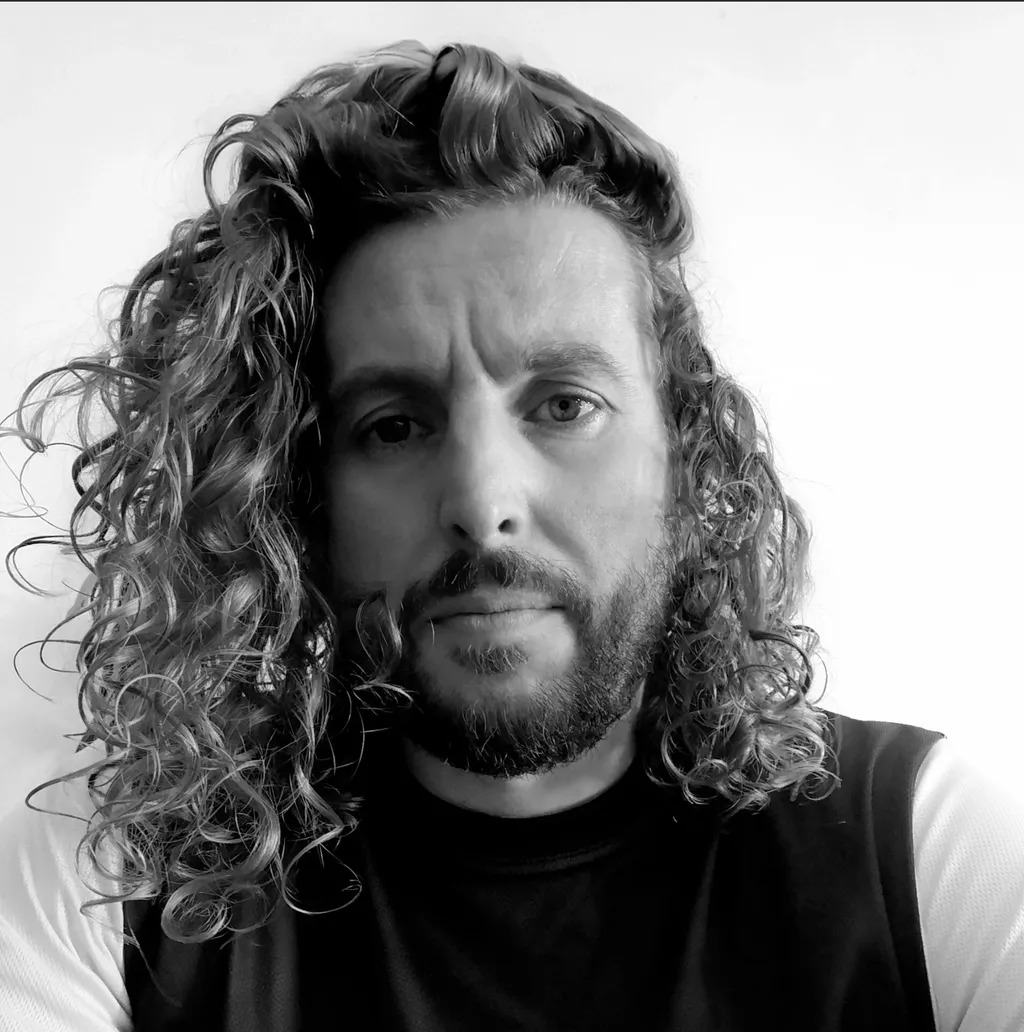 Black and white portrait of man with shoulder-length curly hair and full beard, wearing dark sleeveless top against light background.