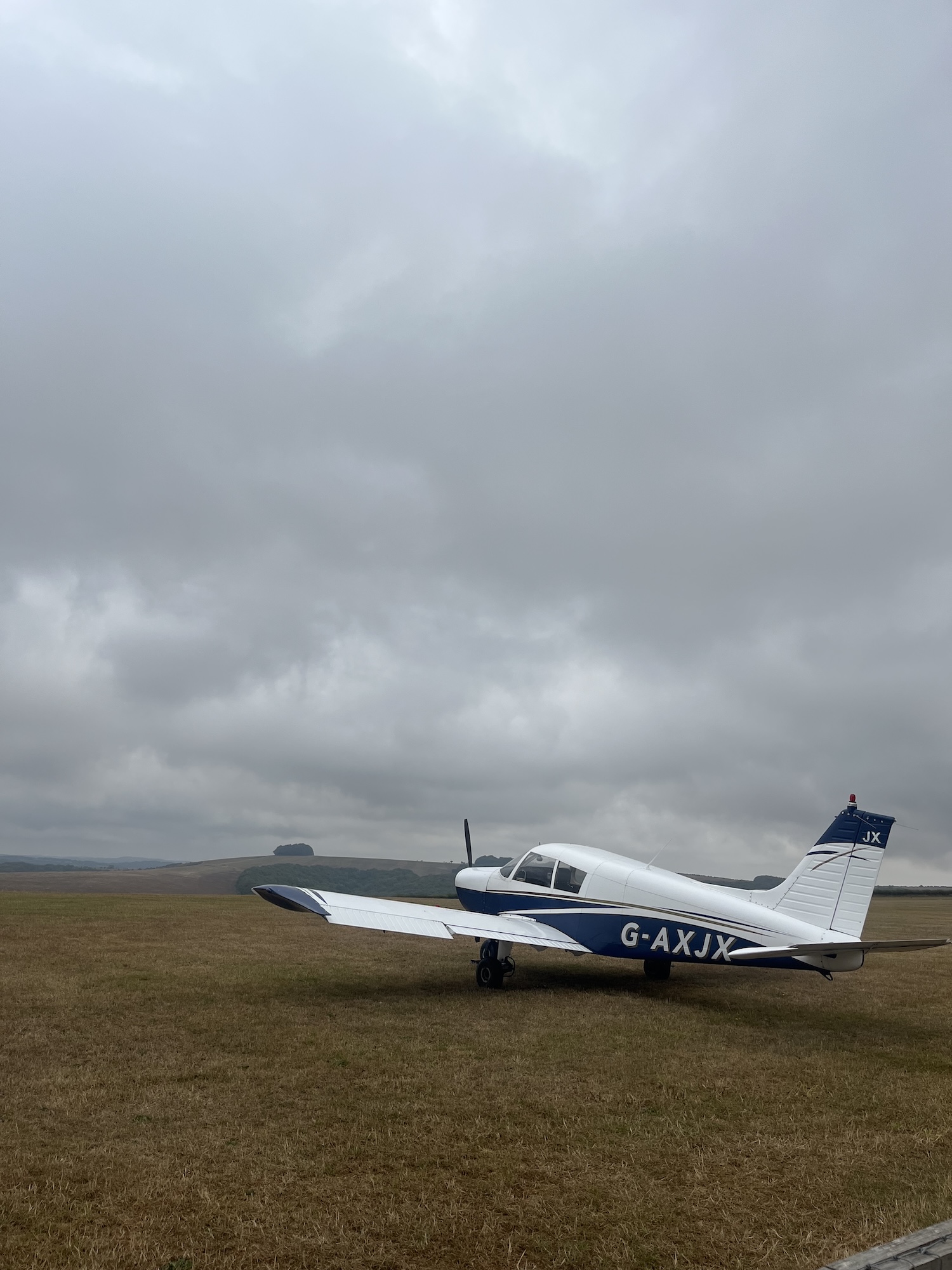A small white and blue propeller aircraft sits on a grassy airfield under heavy grey clouds.