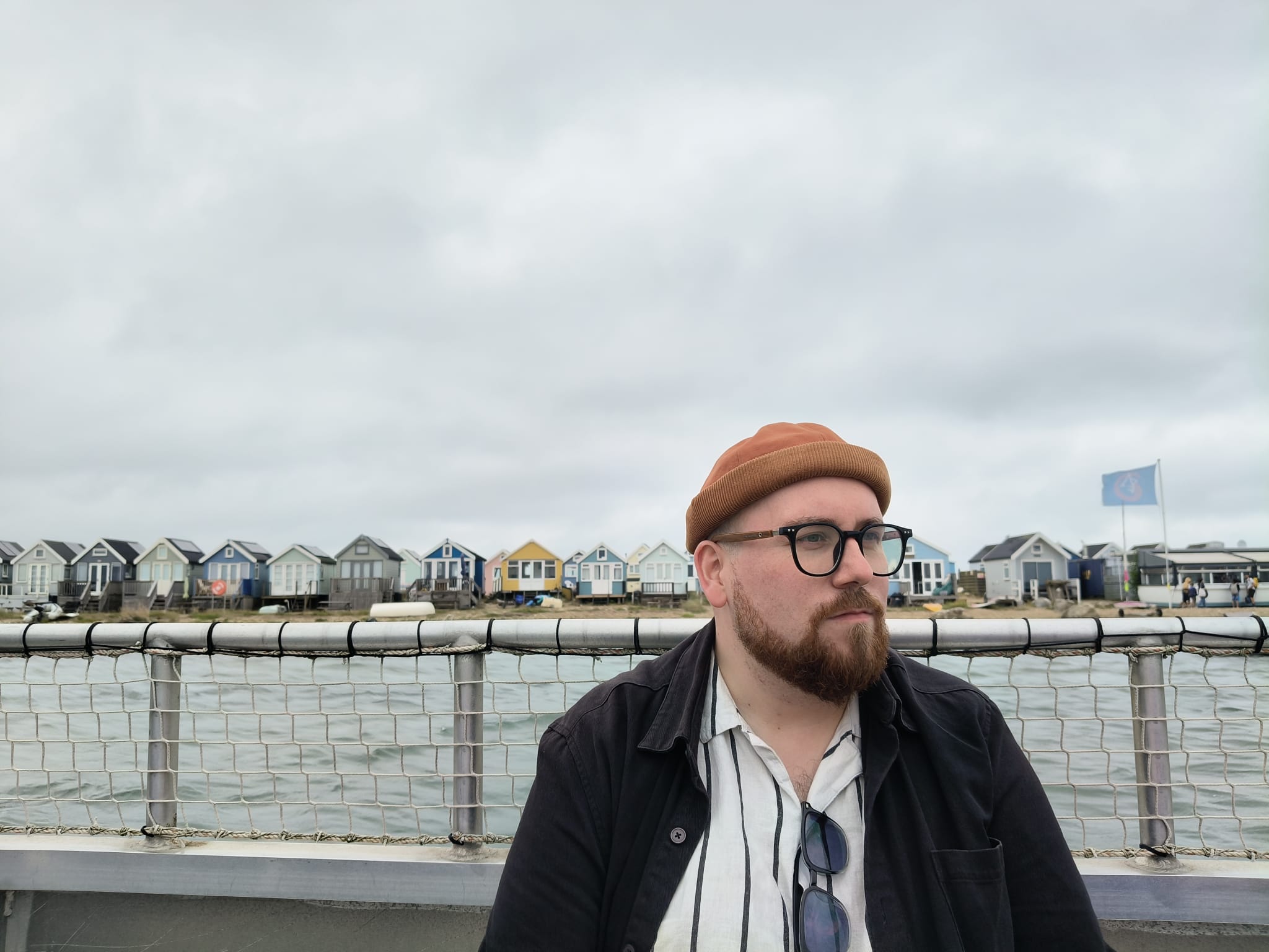A person stands by a seaside railing, with colourful beach huts and water behind him under a cloudy sky.