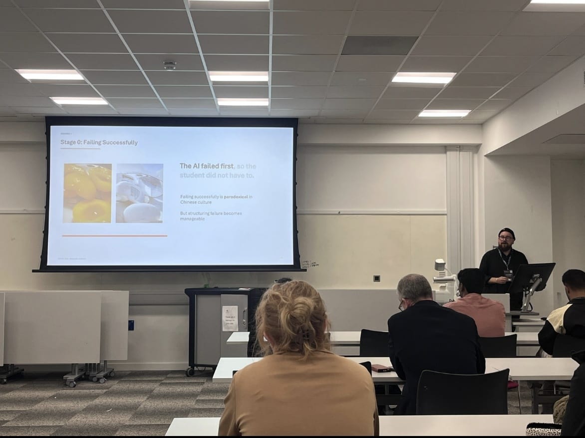 A lecturer stands beside a projector screen in a classroom, with seated attendees facing the front and desks arranged in rows.