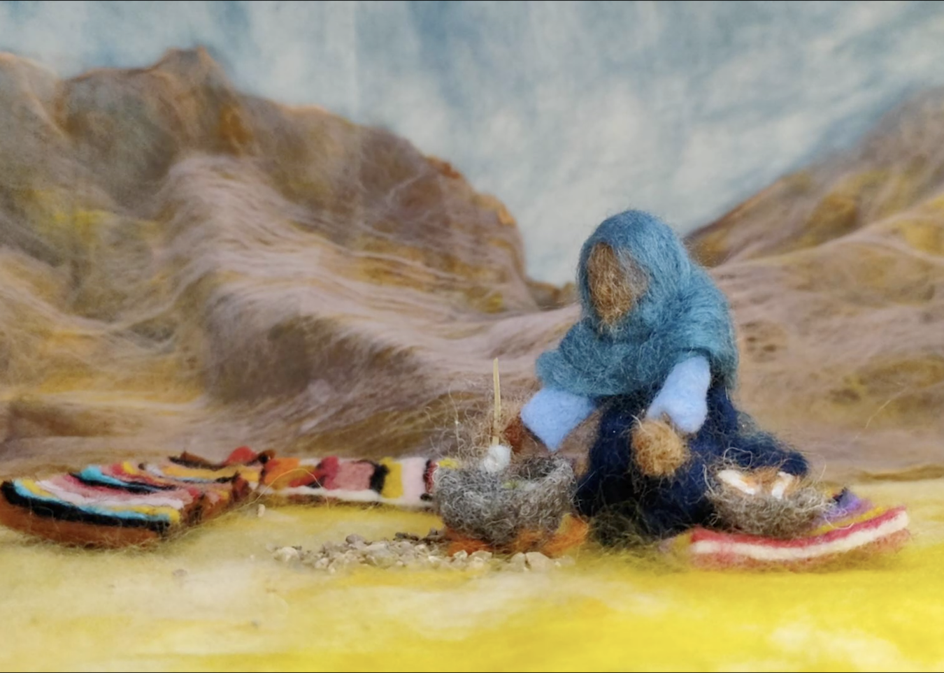 Person in blue hooded clothing sits amongst colourful objects on yellow ground. Brown and beige mountainous landscape behind under pale sky.
