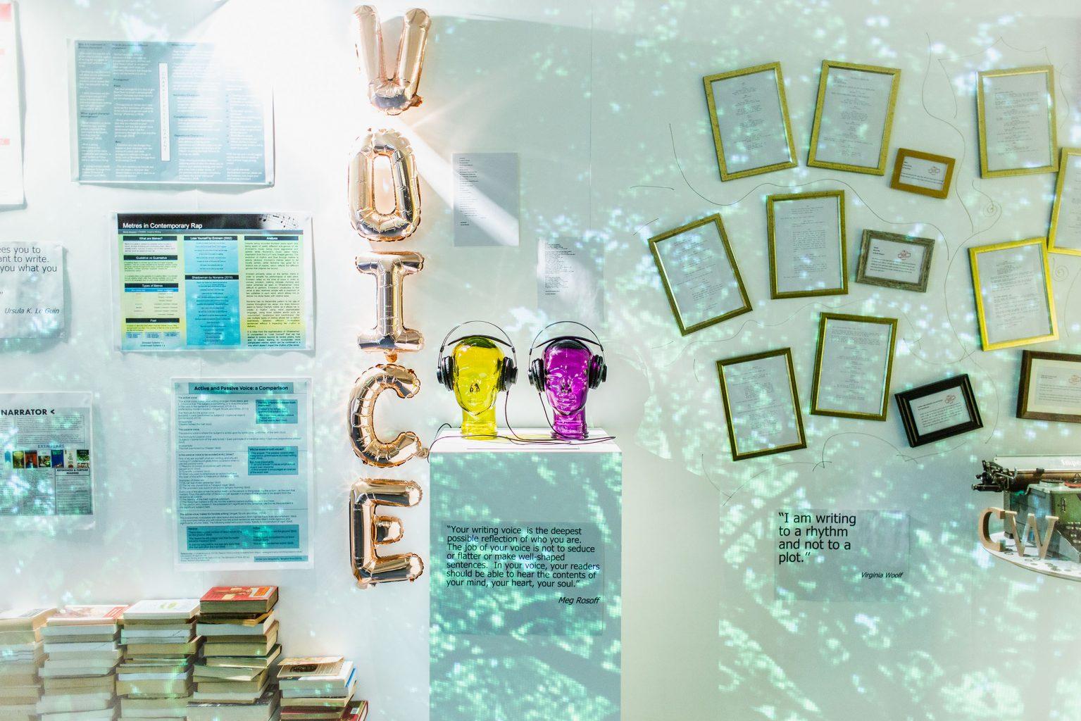 A gallery wall with documents and framed pages on the walls. Two mannequin heads with headphones on stand on a pedestal in the centre. Five balloons arranged vertically spell the word 'Voice'.