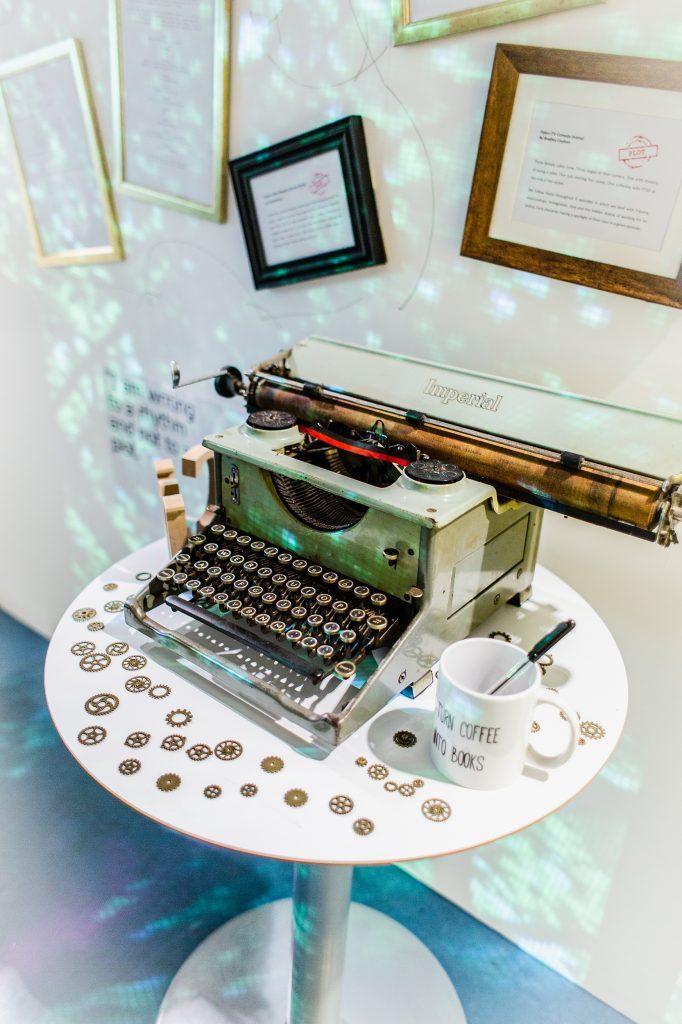 A typewriter standing on a small round table. There are tilted picture frames on the walls behind it.