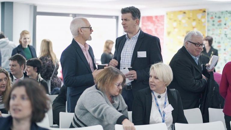 Guests at an event stand and talk. Other people are sitting and talking.