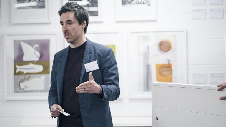 A person delivers a talk in front of several framed pictures, which hang on the wall in the background