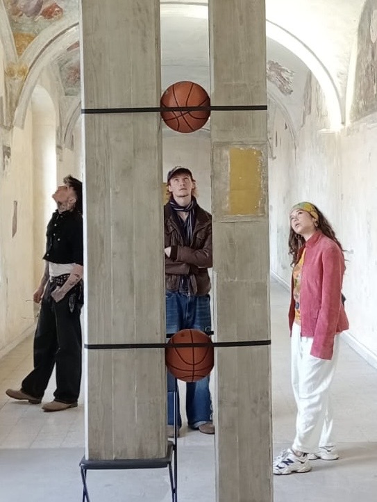 Three people stand among tall concrete pillars in a bright vaulted corridor, with two basketballs fixed between the pillars at chest and head height.