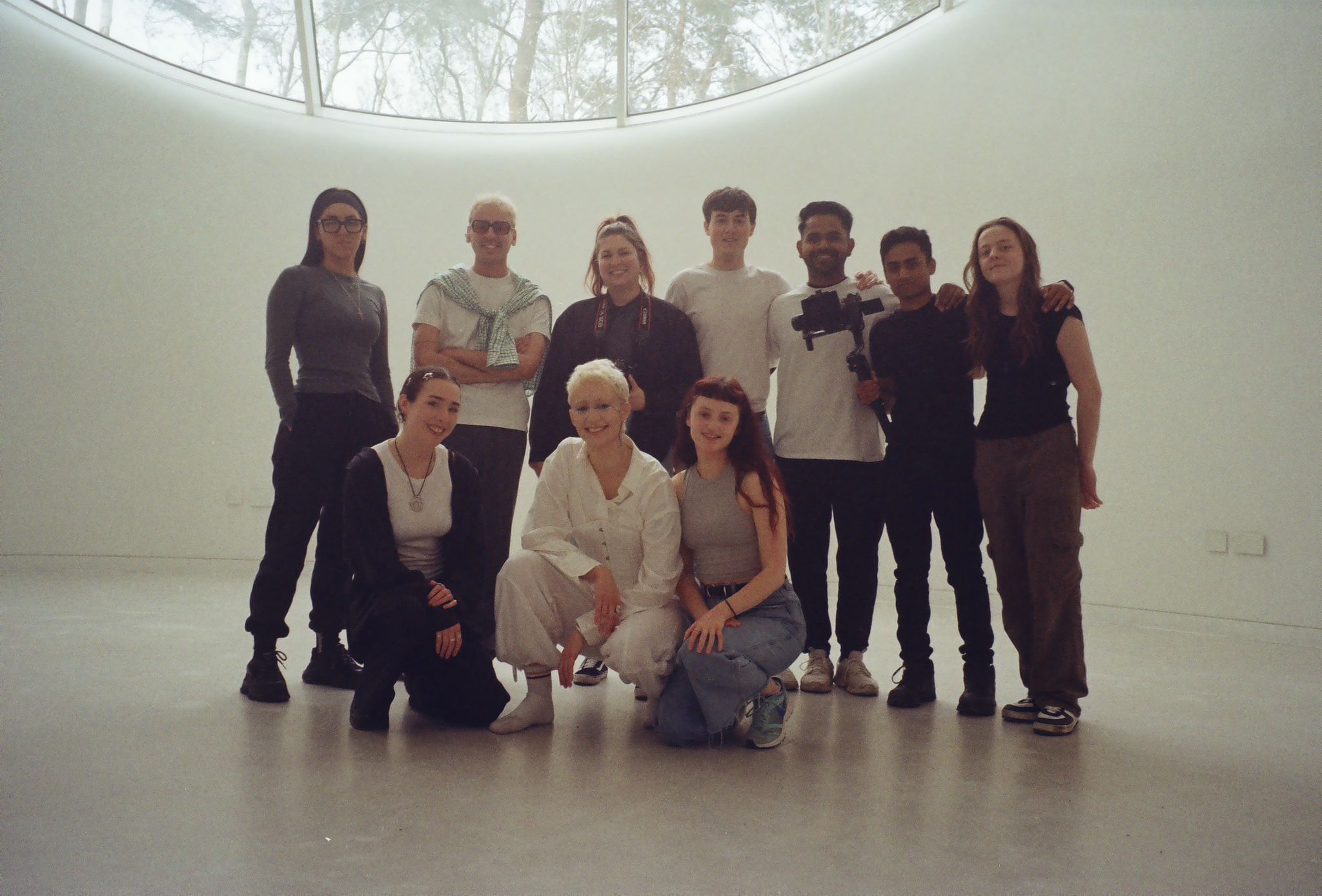 Group of ten people posing in white circular room with skylight overhead showing trees above.