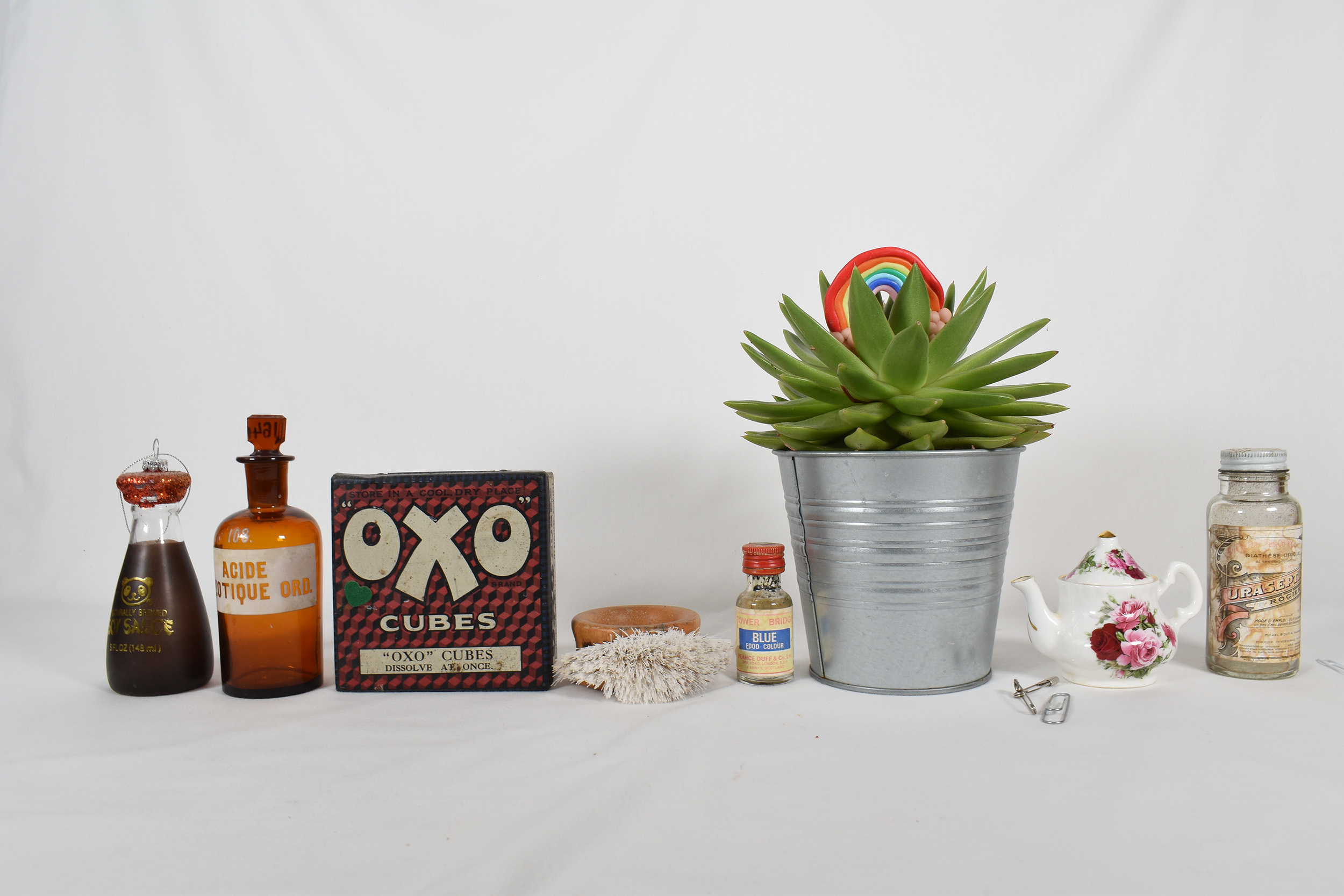 Collection of vintage objects organised from dark to light. Objects include soy sauce bottle, oxo cubes box, succulent plant in a silver pot and a white tea pot with pink floral patterns