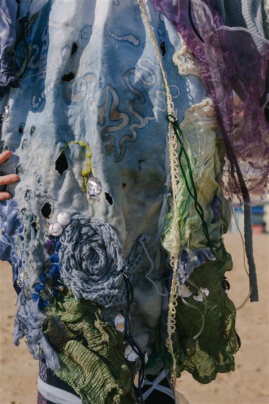 A person wearing a pollution-inspired dress on Bournemouth beach, blue, green and white with patterns and embroidery, close-up.