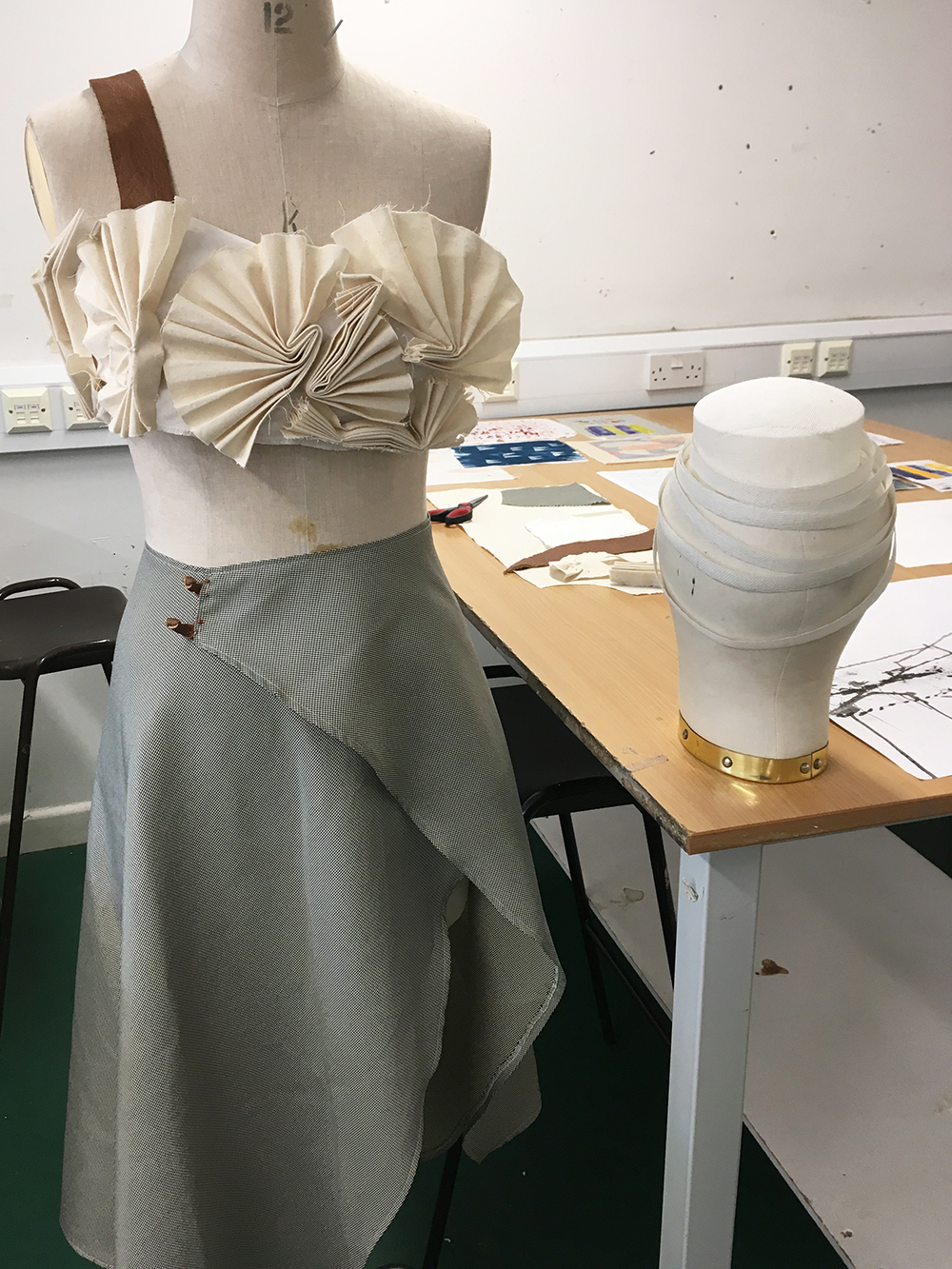 Dress form wearing cream pleated top and sage green wrap skirt in design studio with white ceramic vase on wooden table.